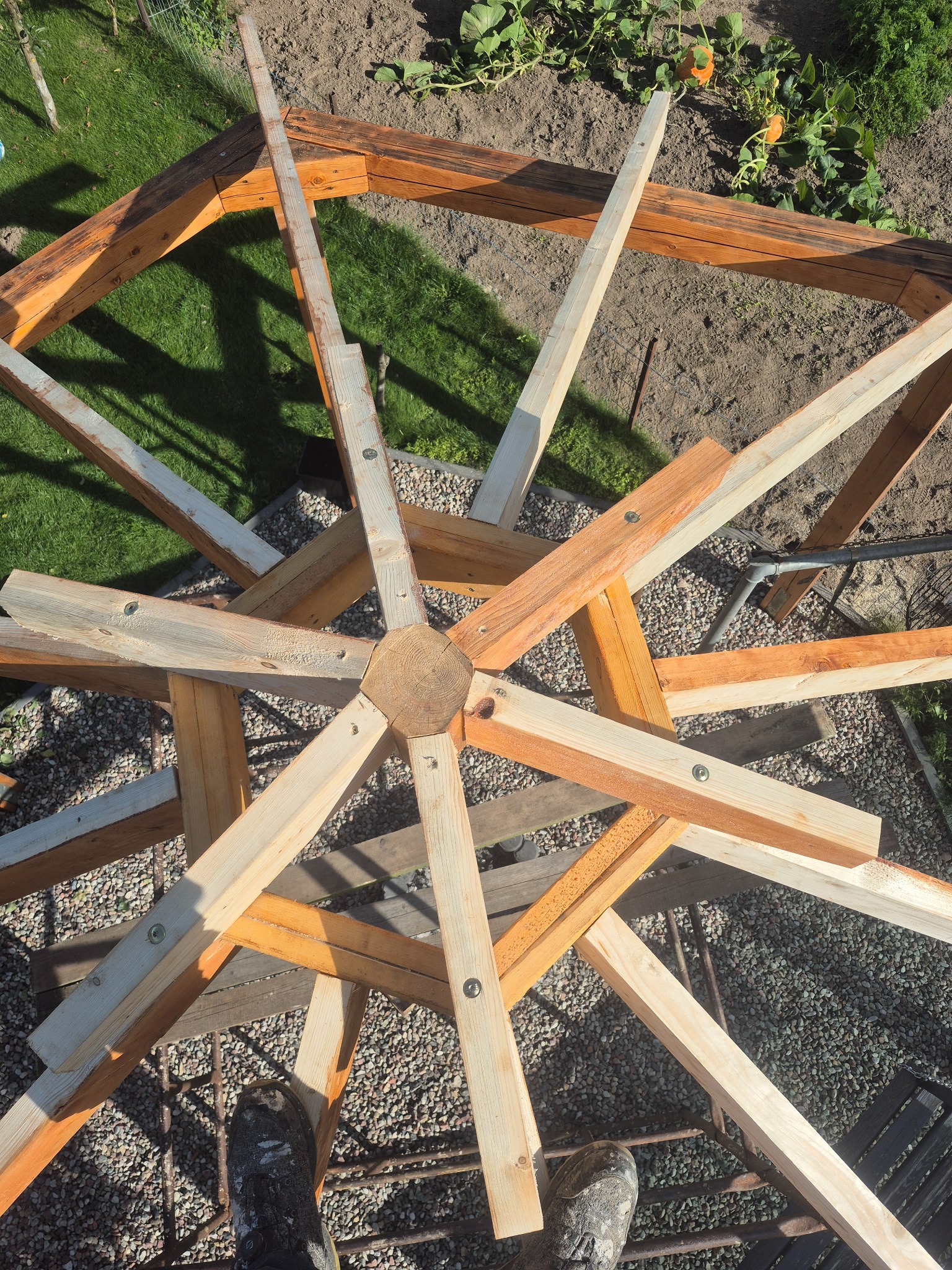 Wiązka drewniana w trakcie montażu, widok z góry na konstrukcję z jasnego drewna, złączona śrubami, na tle ziemi i zieleni. Elementy więźby dachowej.