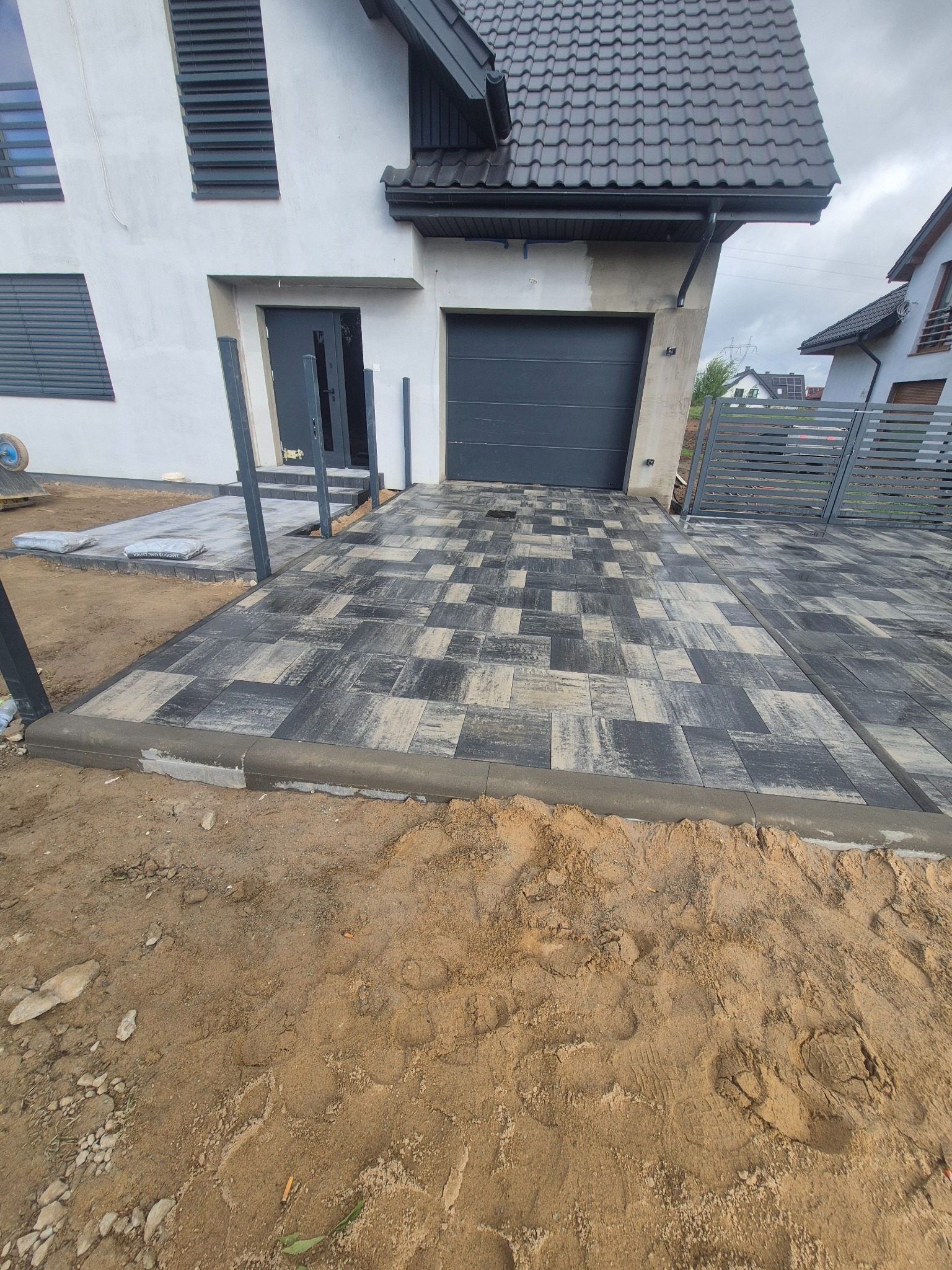 Ułożona kostka brukowa na podjeździe w Szczytnie, widok na dom z szarą bramą garażową i częściowo ułożony chodnik. Widoczne worki z zaprawą.