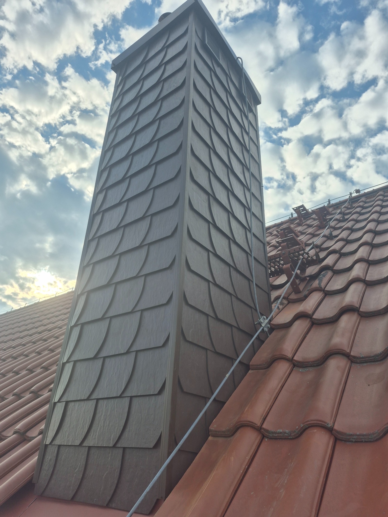 Oryginalne wykończenie komina łupkiem imitującym rybią łuskę, na tle czerwonej dachówki i błękitnego nieba z chmurami. Widoczne zabezpieczenia przeciwśniegowe.