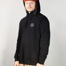 Mężczyzna w czarnej bluzie z kapturem i pomarańczowej czapce typu beanie prezentuje odzież z delikatnym haftowanym wzorem na piersi.