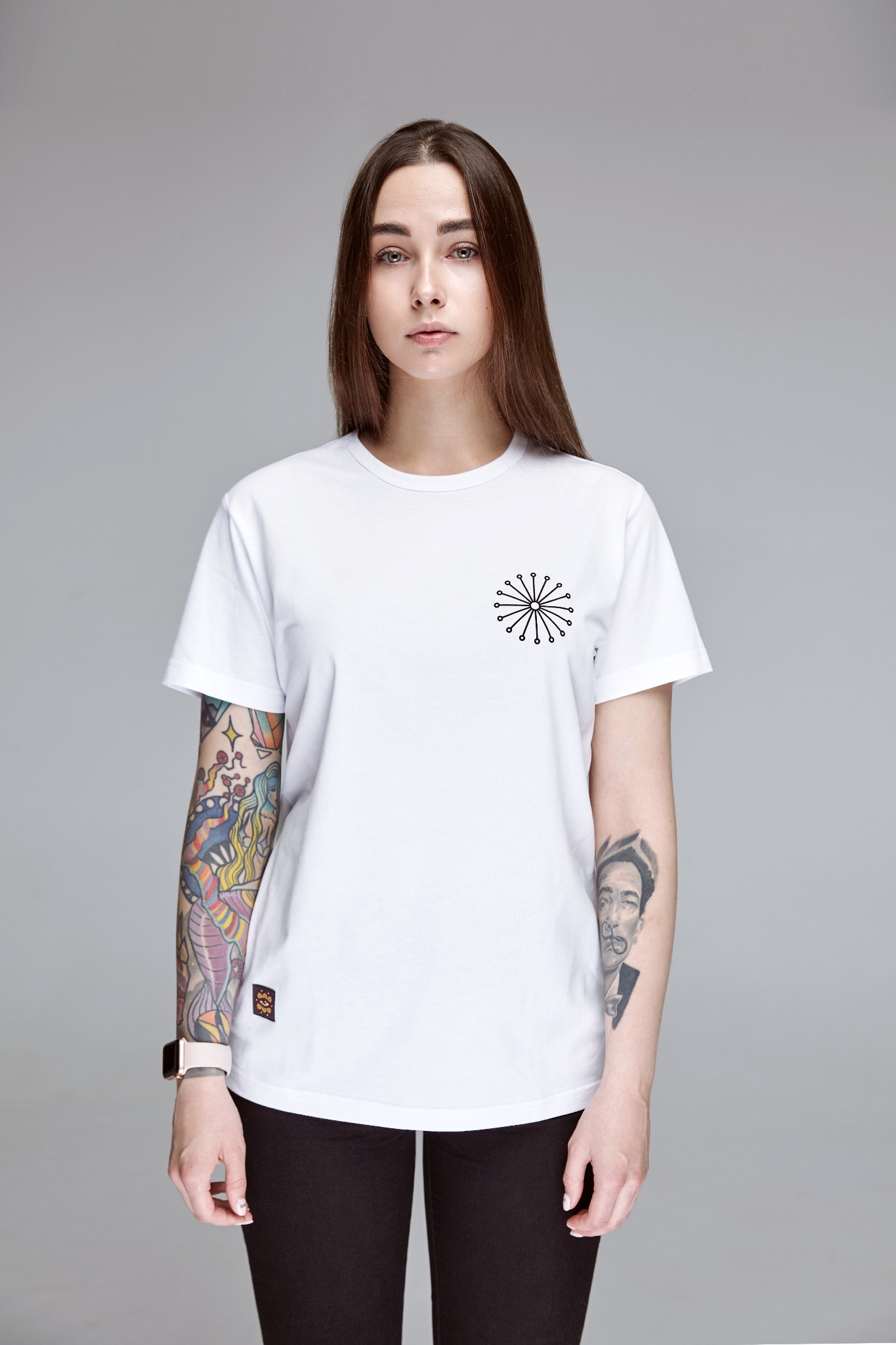 Portret kobiety w białej koszulce z minimalistycznym nadrukiem, widoczne tatuaże na ramionach na tle szarego studia.