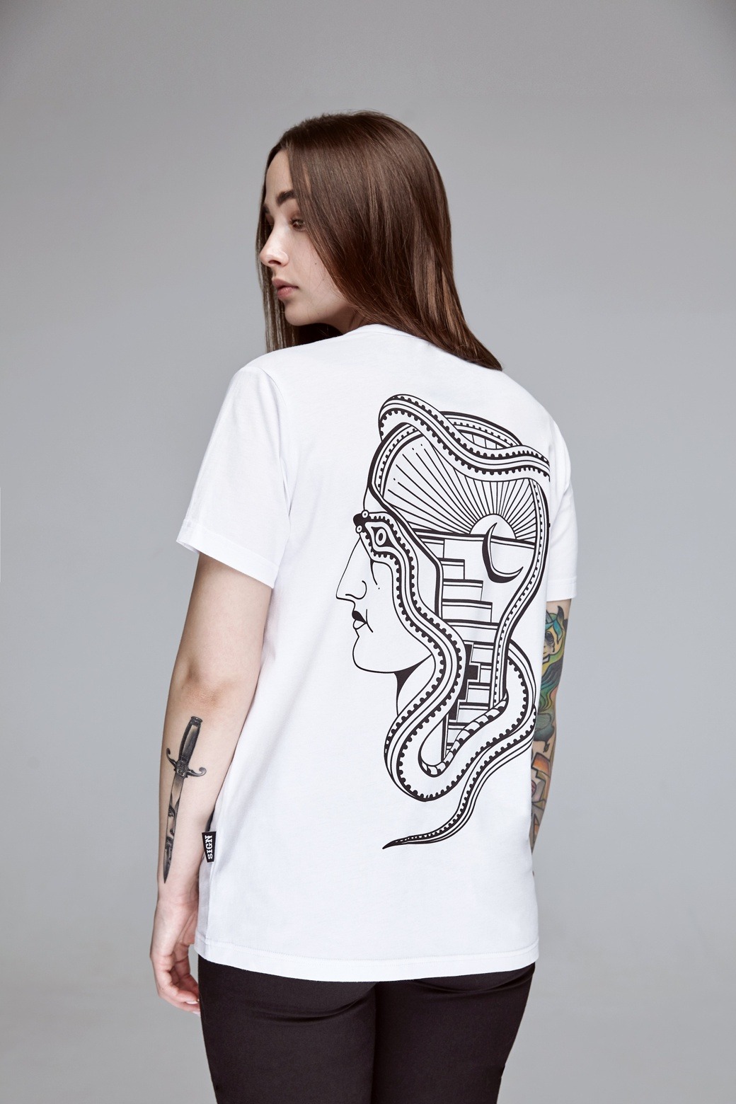 Kobieta w białej koszulce z czarnym nadrukiem przedstawiającym profil twarzy z wężami i schodami na plecach, widok z tyłu, tatuaże na ramionach, Łomianki.