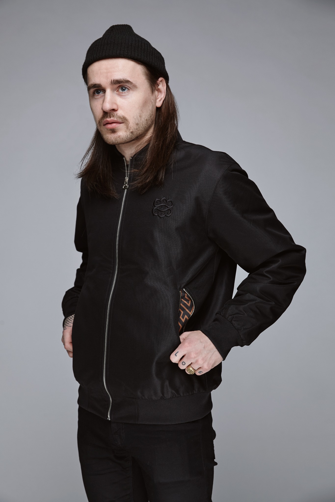 Mężczyzna w czarnej kurtce typu bomber z haftowanym logo na piersi, czarnej czapce i długimi włosami pozuje na szarym tle.