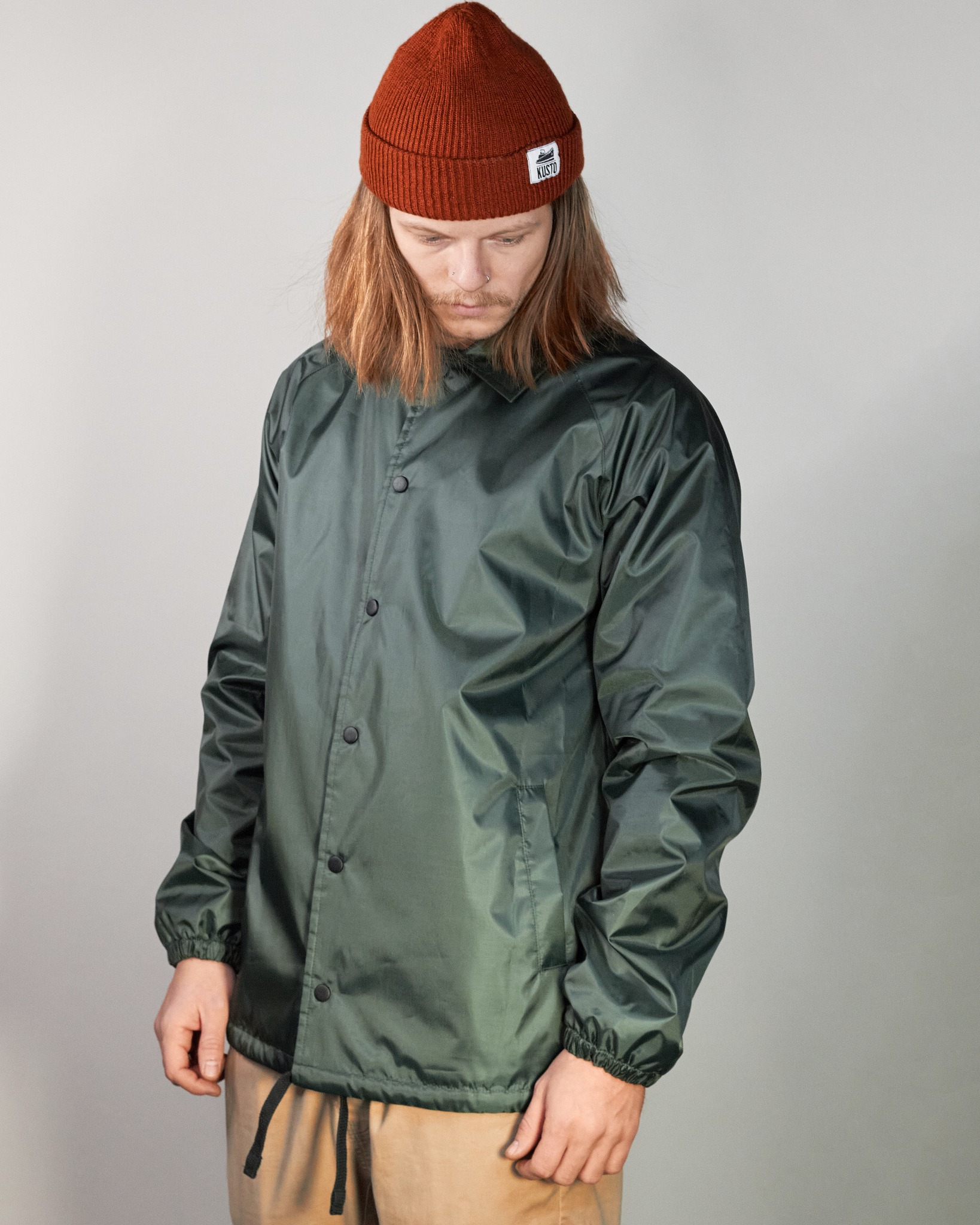 Mężczyzna w brązowej czapce i zielonej kurtce typu coach z czarnymi zatrzaskami, prezentujący produkt na neutralnym tle.