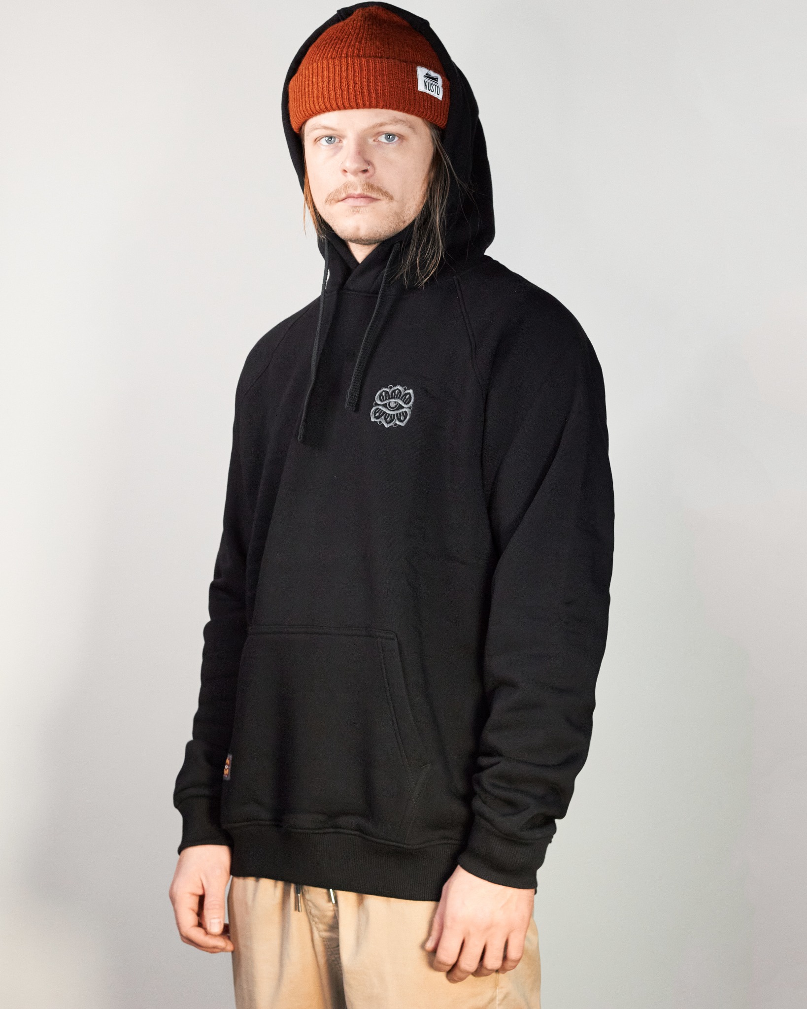 Mężczyzna w czarnej bluzie z kapturem i pomarańczowej czapce typu beanie prezentuje odzież z delikatnym haftowanym wzorem na piersi.
