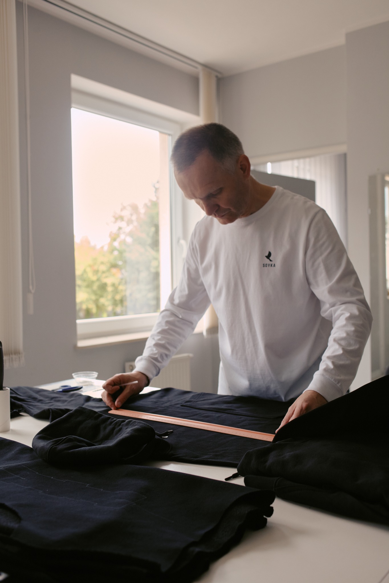 Krawiec w białej bluzie z logo SOYKA mierzy ciemny materiał na stole przy oknie, używając drewnianej linijki i kredy krawieckiej, w tle widoczne inne elementy odzieży.