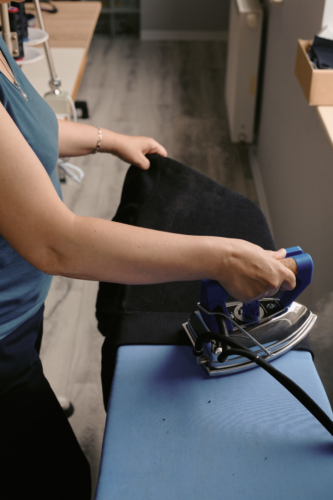 Prasowanie ciemnej bluzy na desce parowym żelazkiem z niebieskim uchwytem; widoczna para, ramię kobiety w niebieskiej koszulce i srebrna bransoletka.