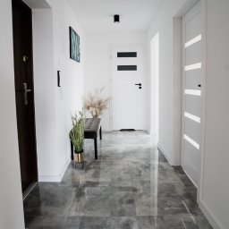 Antal Polska Konrad Kucharczyk - Nowoczesny korytarz z szarymi, polerowanymi płytkami podłogowymi, białymi ścianami, ciemnymi drzwiami wejściowymi i dwiema parami drzwi wewnętrznych, oświetlony czarnymi lampami sufitowymi.
