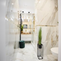 Antal Polska Konrad Kucharczyk - Elegancka łazienka z marmurowymi płytkami, złotymi akcentami i minimalistycznym wystrojem, obejmująca wiszącą umywalkę, kabinę prysznicową ze złotymi ramami i dekoracyjną roślinę w czarnej donicy.