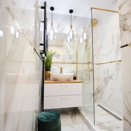 Antal Polska Konrad Kucharczyk - Elegancka łazienka z marmurowymi płytkami, kabiną prysznicową ze złotymi akcentami, owalną umywalką na drewnianym blacie, białymi szafkami z złotymi uchwytami i zielonym pufem.