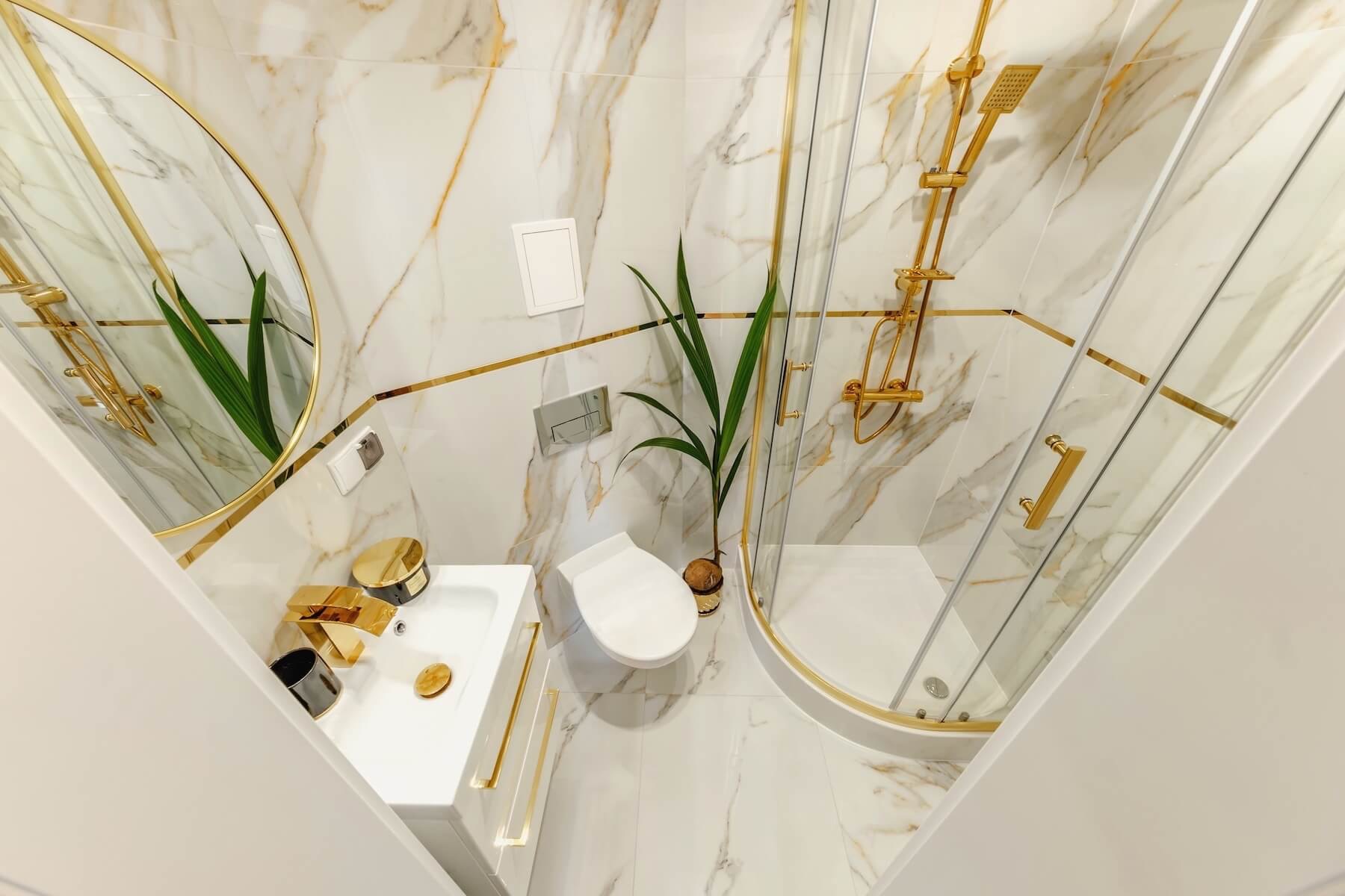 Wykończona łazienka z białymi marmurowymi płytkami z żyłami w kolorze złota, złotą armaturą, kabiną prysznicową typu walk-in i okrągłym lustrem, widok z góry.