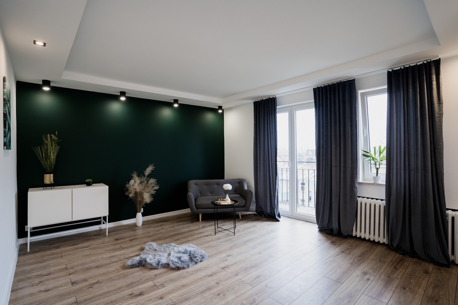 Jasne wnętrze salonu z drewnianą podłogą, ciemnozieloną ścianą akcentującą z punktowym oświetleniem, szarą sofą, białymi meblami oraz balkonem z szarymi zasłonami.