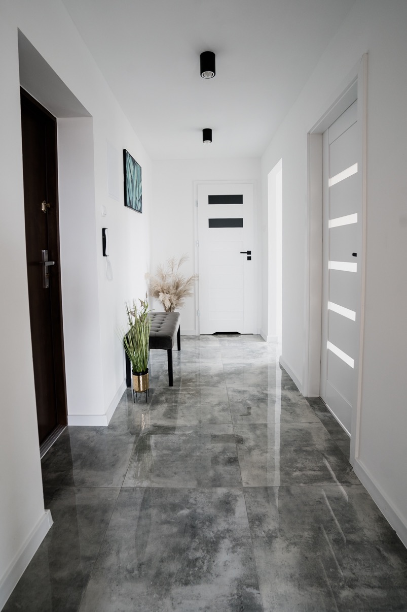 Nowoczesny korytarz z szarymi, polerowanymi płytkami podłogowymi, białymi ścianami, ciemnymi drzwiami wejściowymi i dwiema parami drzwi wewnętrznych, oświetlony czarnymi lampami sufitowymi.
