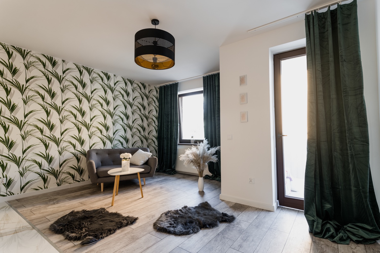 Nowoczesny salon z tapetą w liście, szarą sofą, okrągłym stolikiem kawowym i czarnym ażurowym żyrandolem z elementami złota.