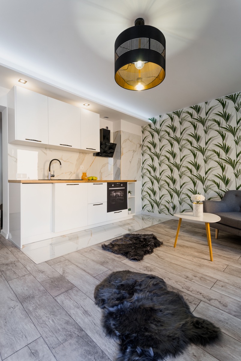 Nowoczesna kuchnia z białymi szafkami, marmurowym backsplashiem i tapetą w liście, oświetlona czarną lampą z ażurowym kloszem i złotym wnętrzem, uzupełniona szarym narożnikiem i dwoma ciemnymi...