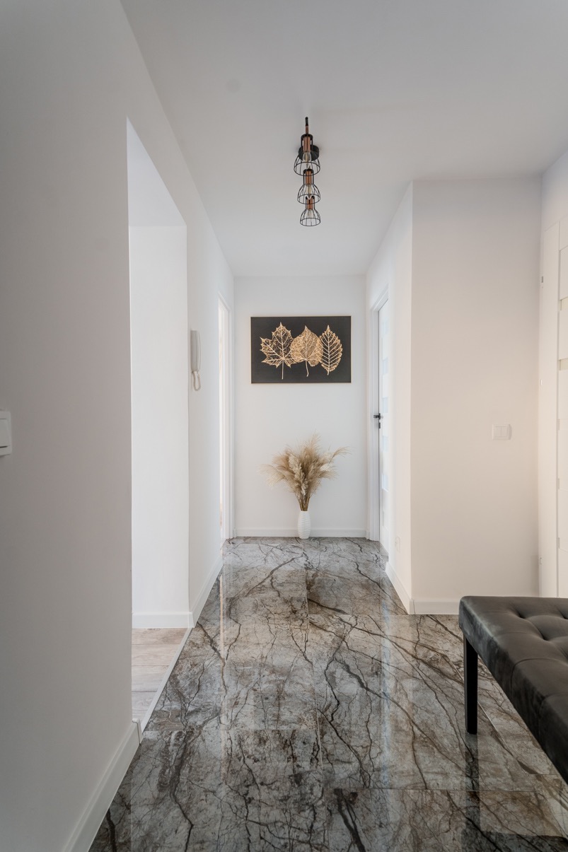 Wykończony korytarz z marmurową posadzką, dekoracją ścienną z motywem liści, bukietem w wazonie i nowoczesną lampą sufitową.