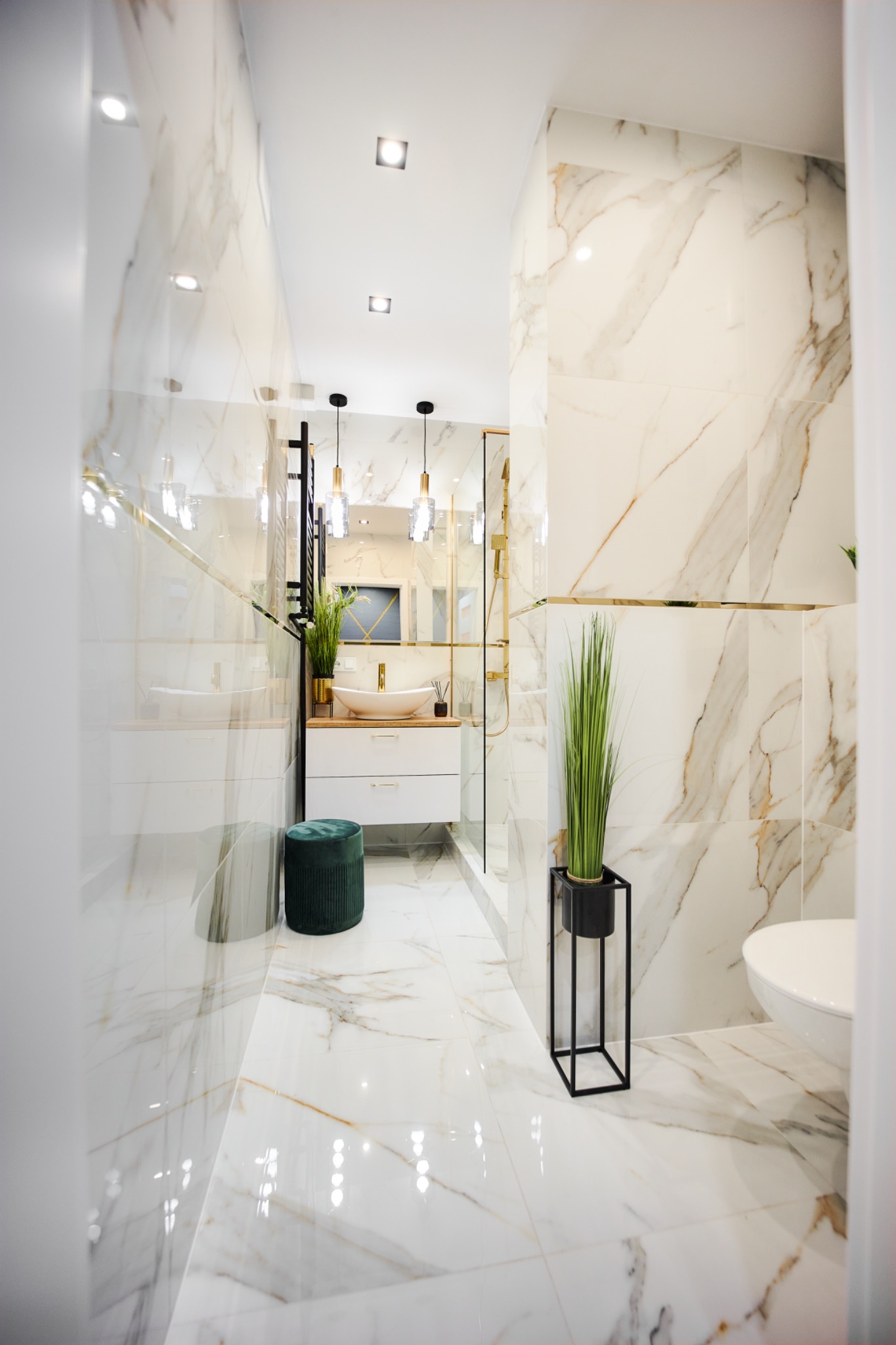 Elegancka łazienka z marmurowymi płytkami, złotymi akcentami i minimalistycznym wystrojem, obejmująca wiszącą umywalkę, kabinę prysznicową ze złotymi ramami i dekoracyjną roślinę w czarnej donicy.