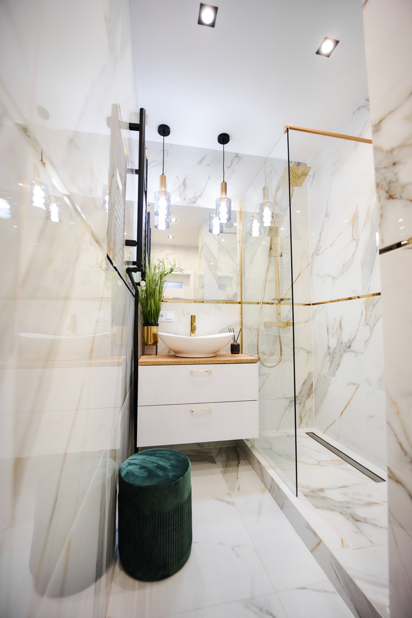 Elegancka łazienka z marmurowymi płytkami, kabiną prysznicową ze złotymi akcentami, owalną umywalką na drewnianym blacie, białymi szafkami z złotymi uchwytami i zielonym pufem.