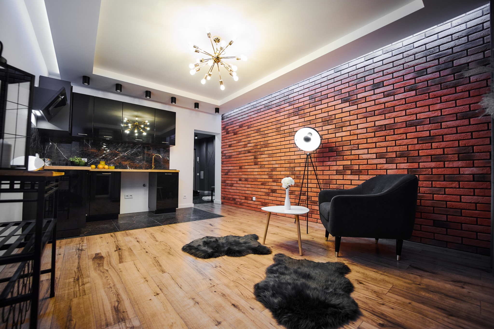 Nowoczesne wnętrze z czarną zabudową kuchenną, marmurowym blatem, czerwoną ścianą z cegły, drewnianą podłogą i designerską lampą sufitową.