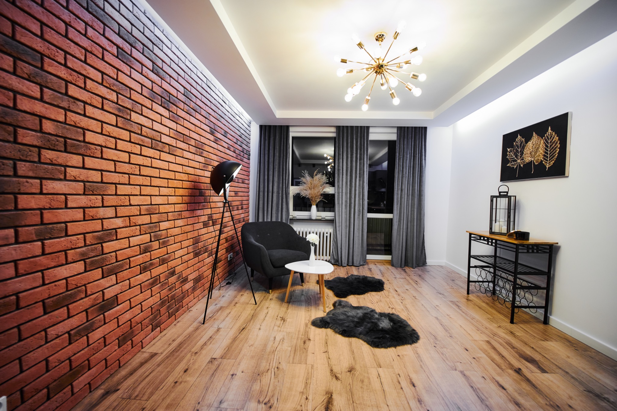 Salon z dekoracyjną ścianą z czerwonej cegły, drewnianą podłogą, czarnym fotelem, okrągłym stolikiem kawowym i lampą podłogową na trójnogu, oświetlony nowoczesną lampą sufitową.