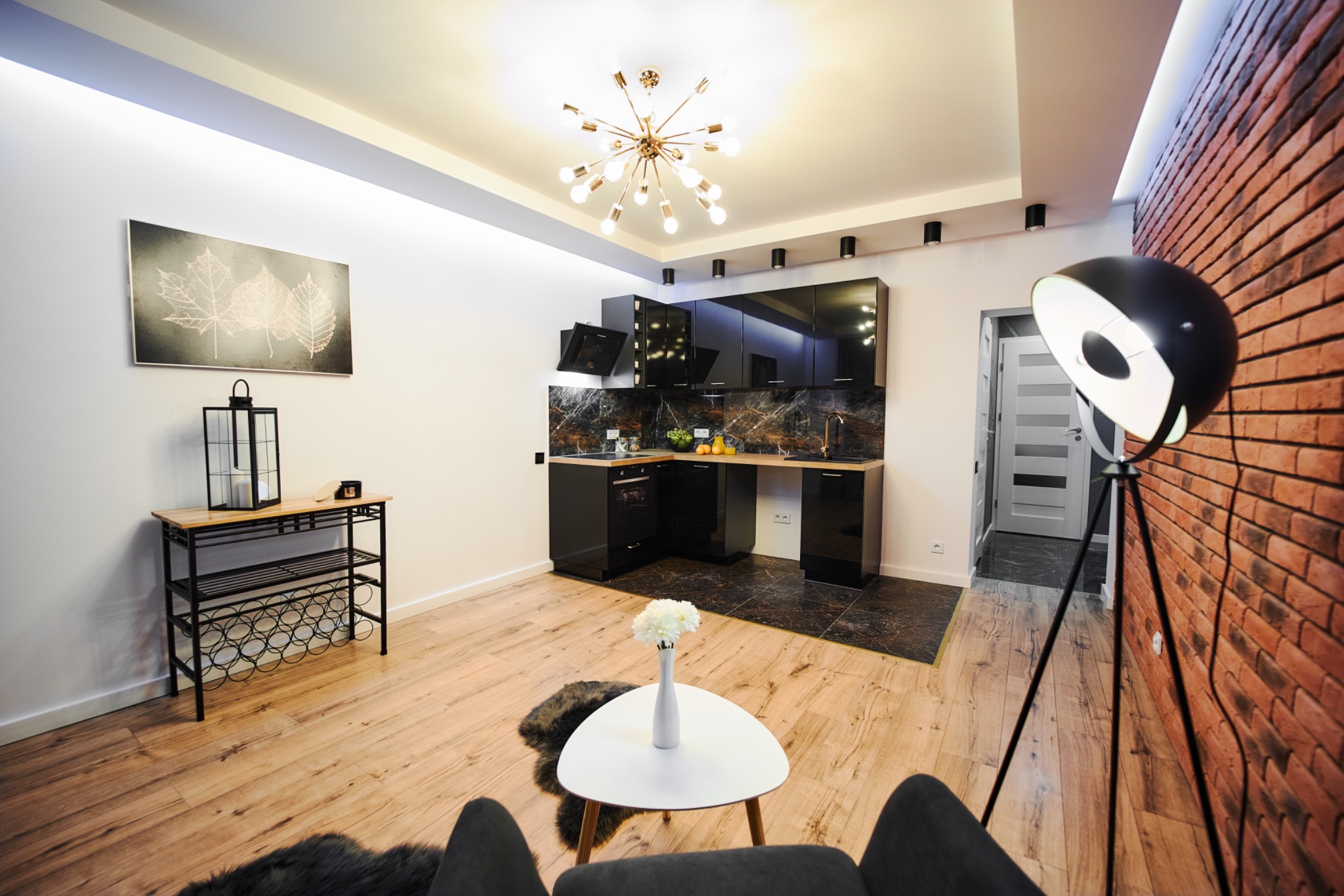 Nowoczesne wnętrze: czarna, błyszcząca zabudowa kuchenna z marmurowym blatem i backsplash, oświetlenie punktowe, drewniana podłoga, ceglana ściana z lampą na trójnogu, stolik kawowy z kwiatami...