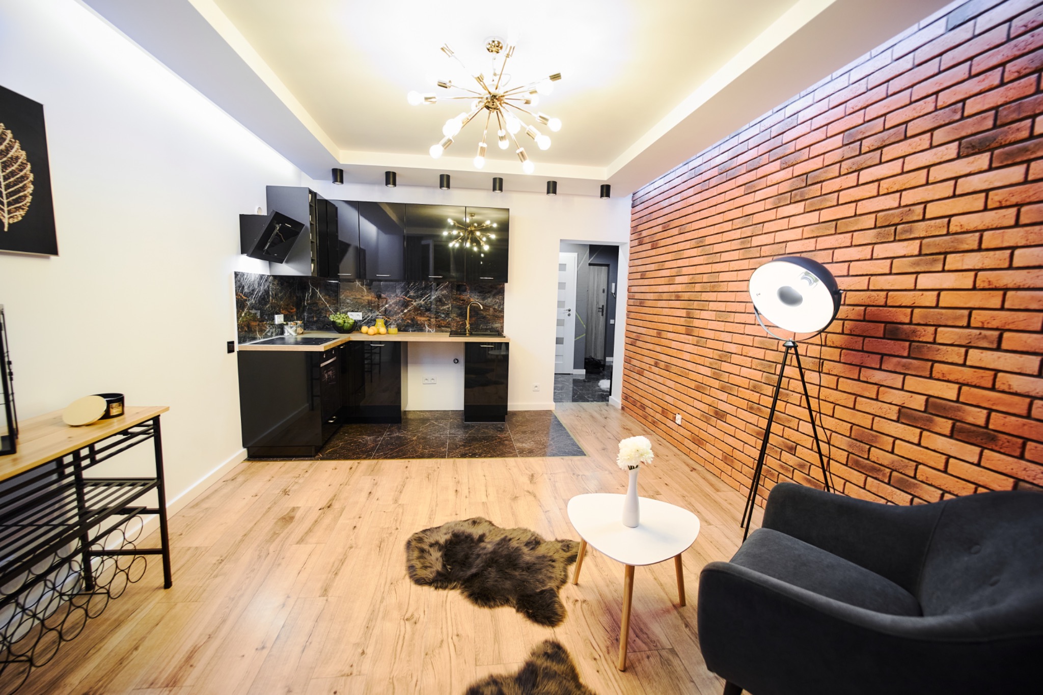 Nowoczesne wnętrze: ceglana ściana, czarna zabudowa kuchenna z marmurowym blatem, designerska lampa, drewniana podłoga i szary fotel tworzą stylową przestrzeń.