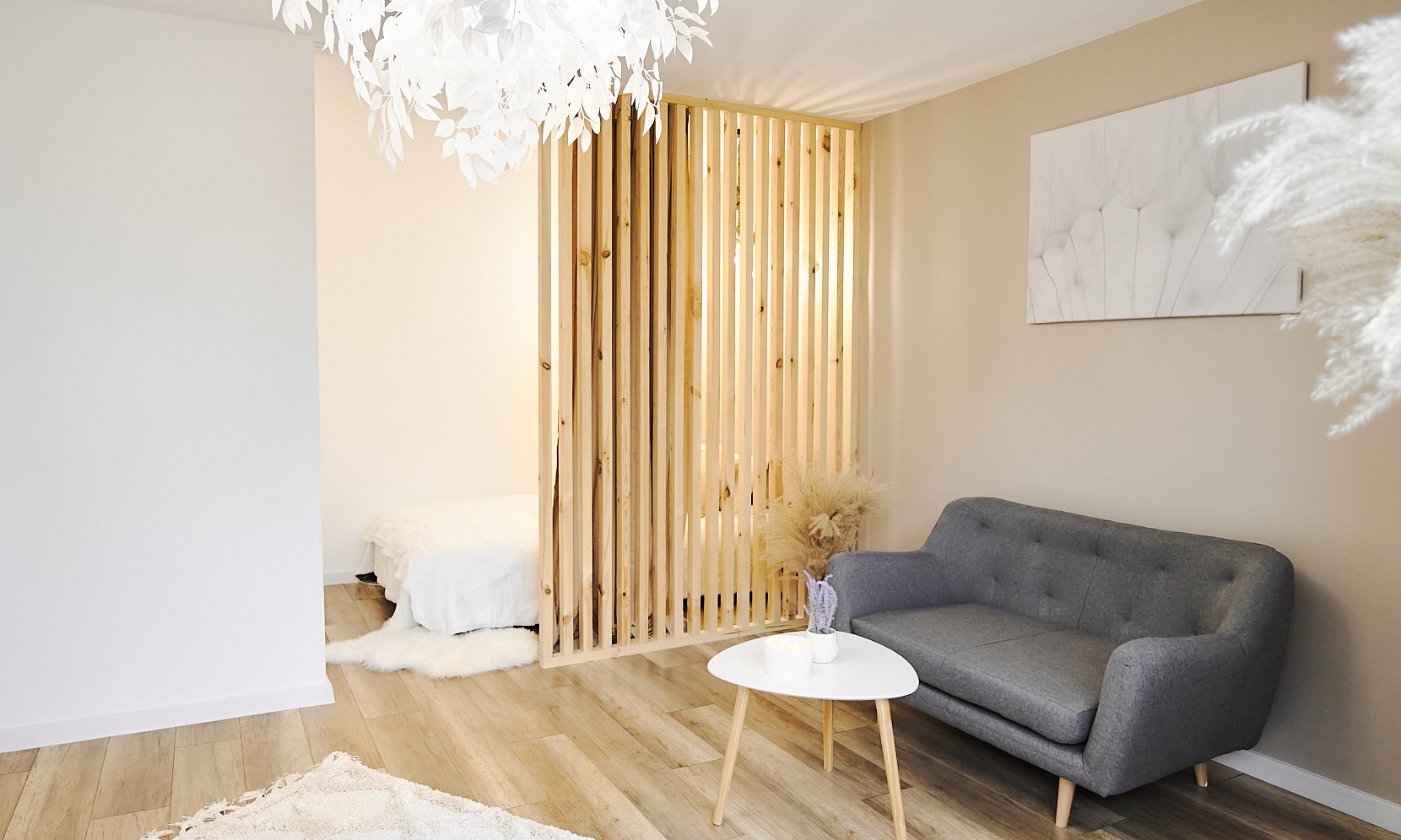 Jasne wnętrze z drewnianą ażurową ścianką działową, szarą sofą, białym stolikiem i dekoracyjną lampą sufitową w kształcie liści.