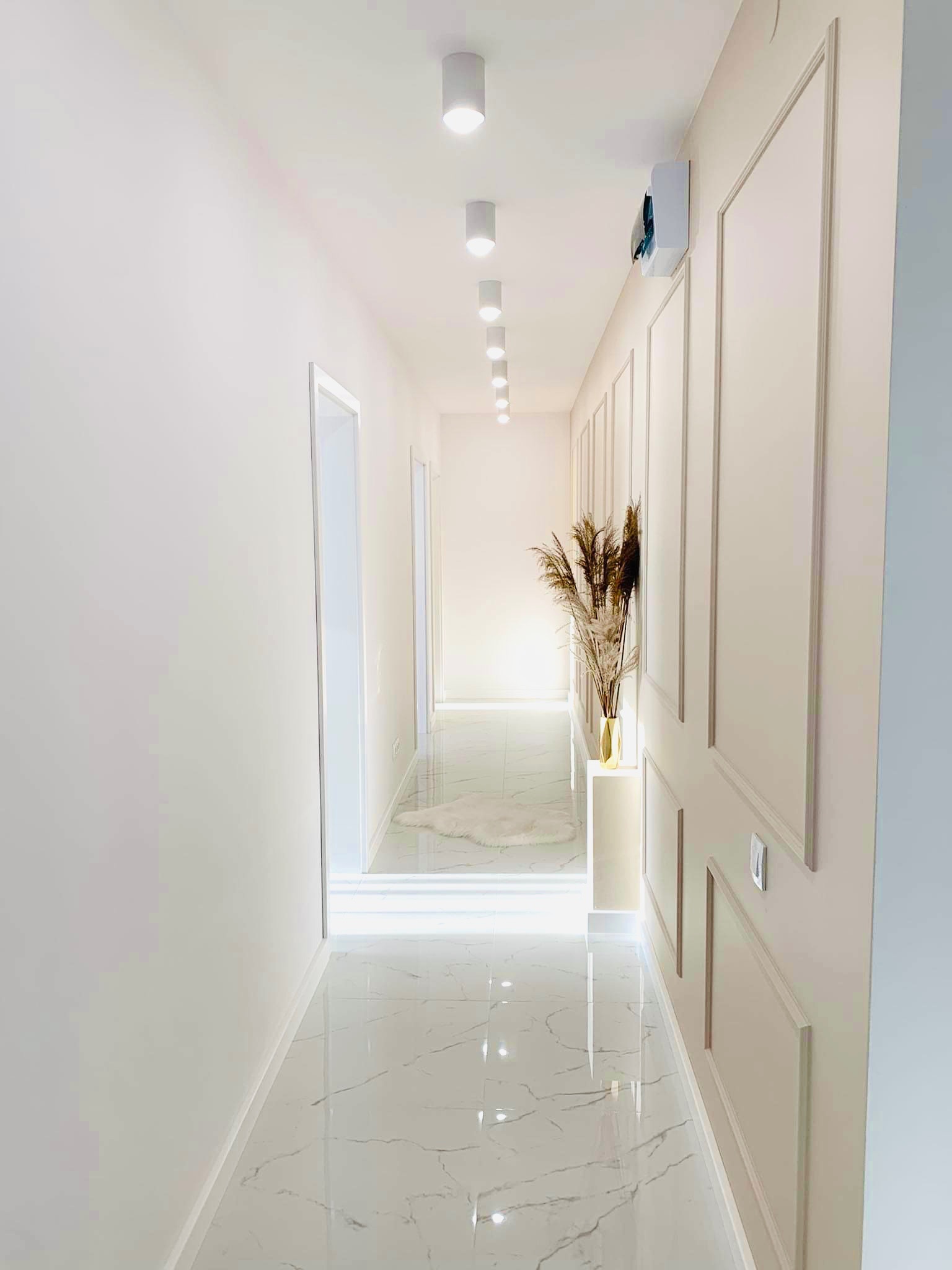 Jasny korytarz z marmurową podłogą, dekoracyjnymi panelami ściennymi, rzędem punktowych lamp sufitowych i wazonem z suszonymi trawami na postumencie.