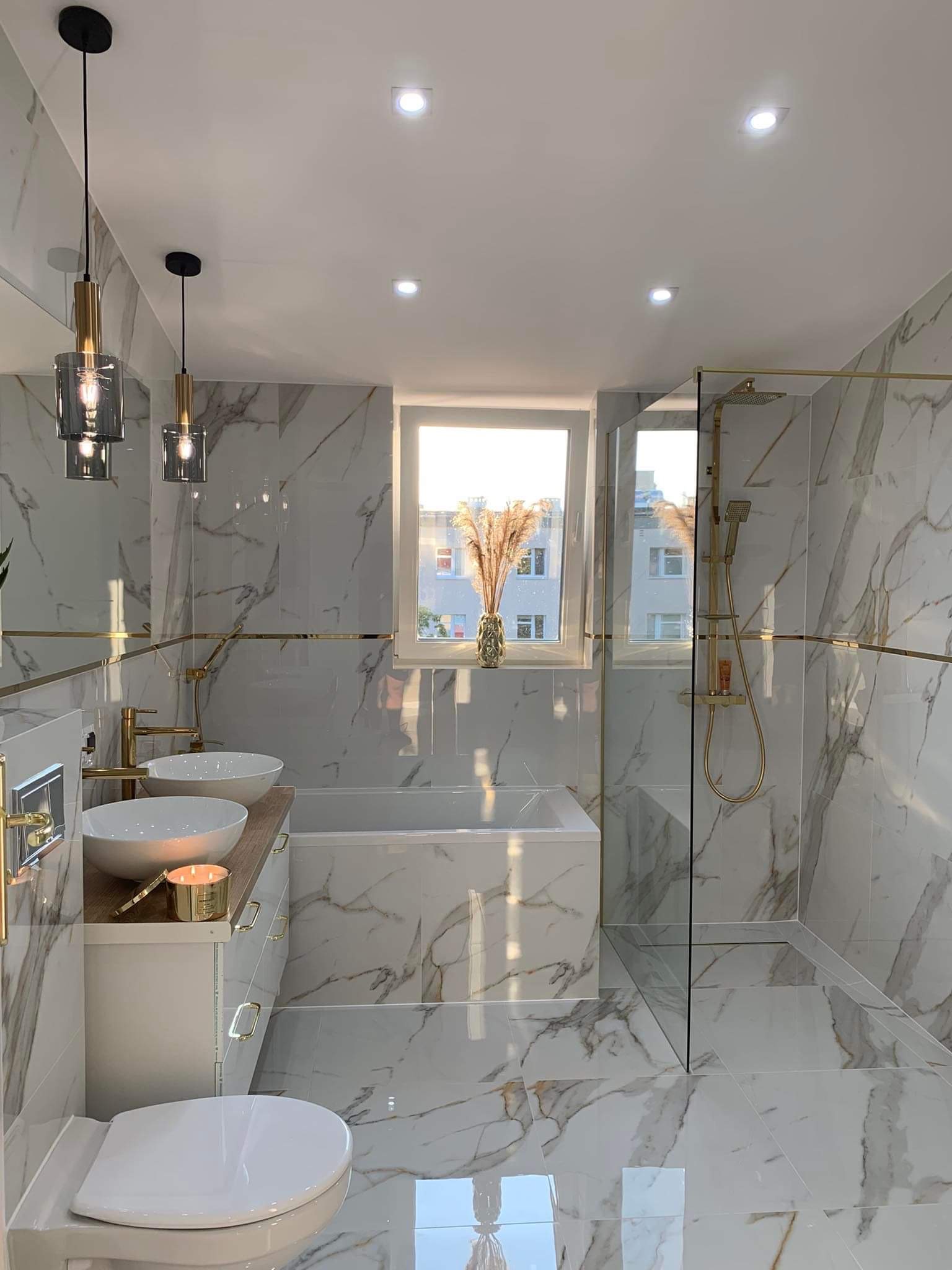 Elegancka łazienka z marmurowymi płytkami, złotymi akcentami w armaturze i oświetleniu, podwójną umywalką i kabiną prysznicową typu walk-in, oświetlona światłem dziennym wpadającym przez okno...