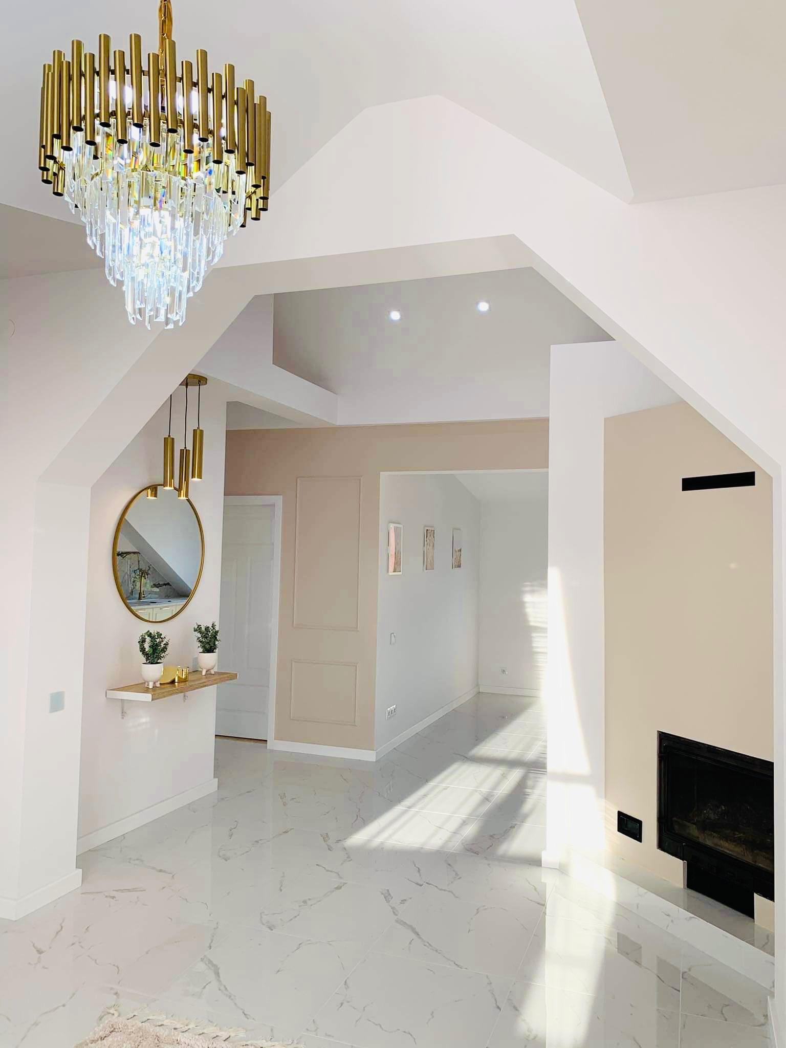 Przestronny, jasny hol z marmurową posadzką, złotym żyrandolem z kryształkami, okrągłym lustrem z złotą ramą i dekoracyjną konsolą z roślinami w doniczkach.