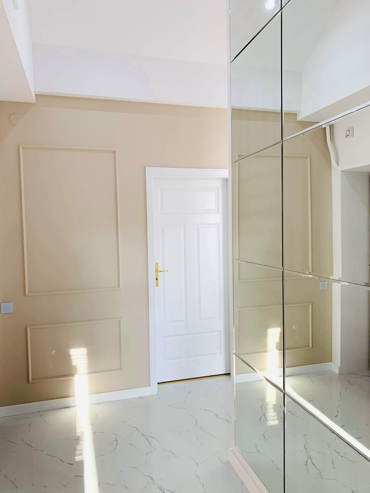Eleganckie wnętrze z beżowymi ścianami z dekoracyjnymi ramami, białe drzwi z złotą klamką, lustrzana szafa w zabudowie i marmurowa podłoga z szarymi żyłami.