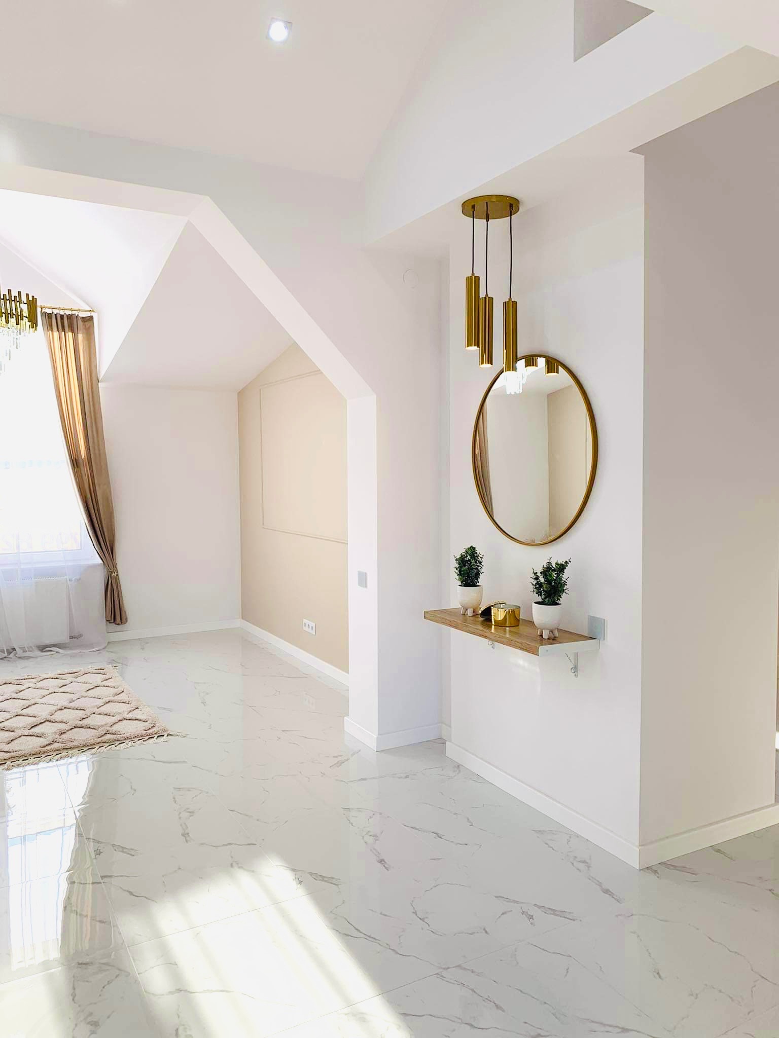 Jasne wnętrze poddasza z marmurową podłogą, okrągłym lustrem w złotej ramie, złotą lampą wiszącą i minimalistyczną półką z dekoracjami.
