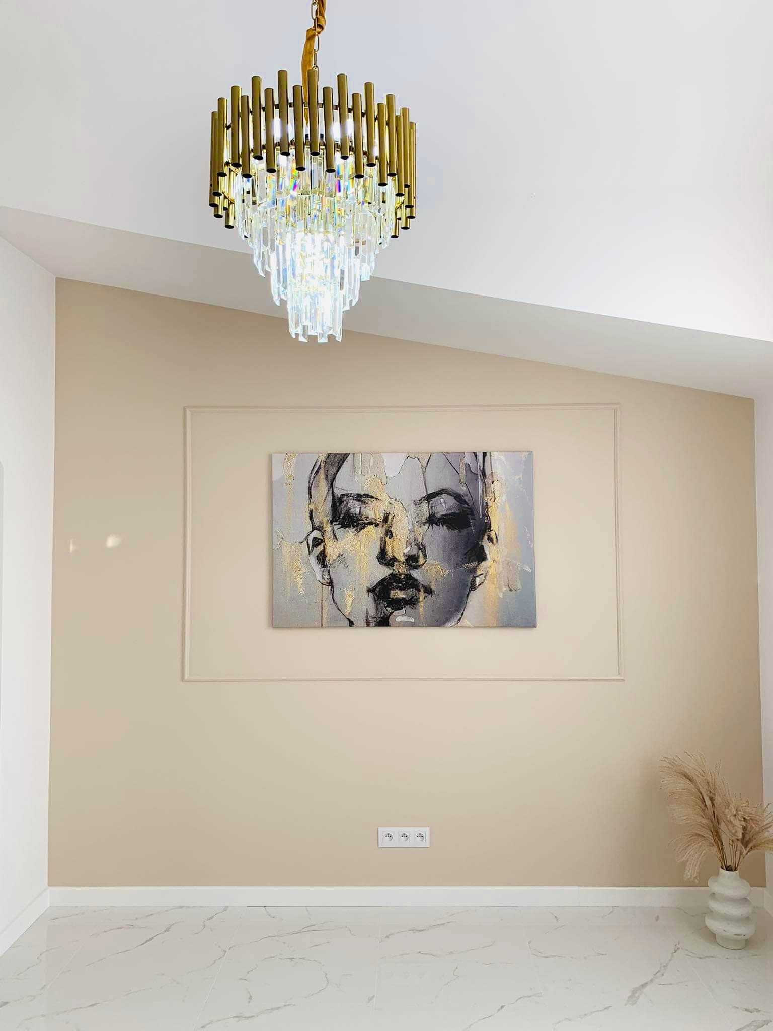 Beżowe wnętrze z marmurową podłogą, obrazem kobiety w ramie i złotym żyrandolem kaskadowym.