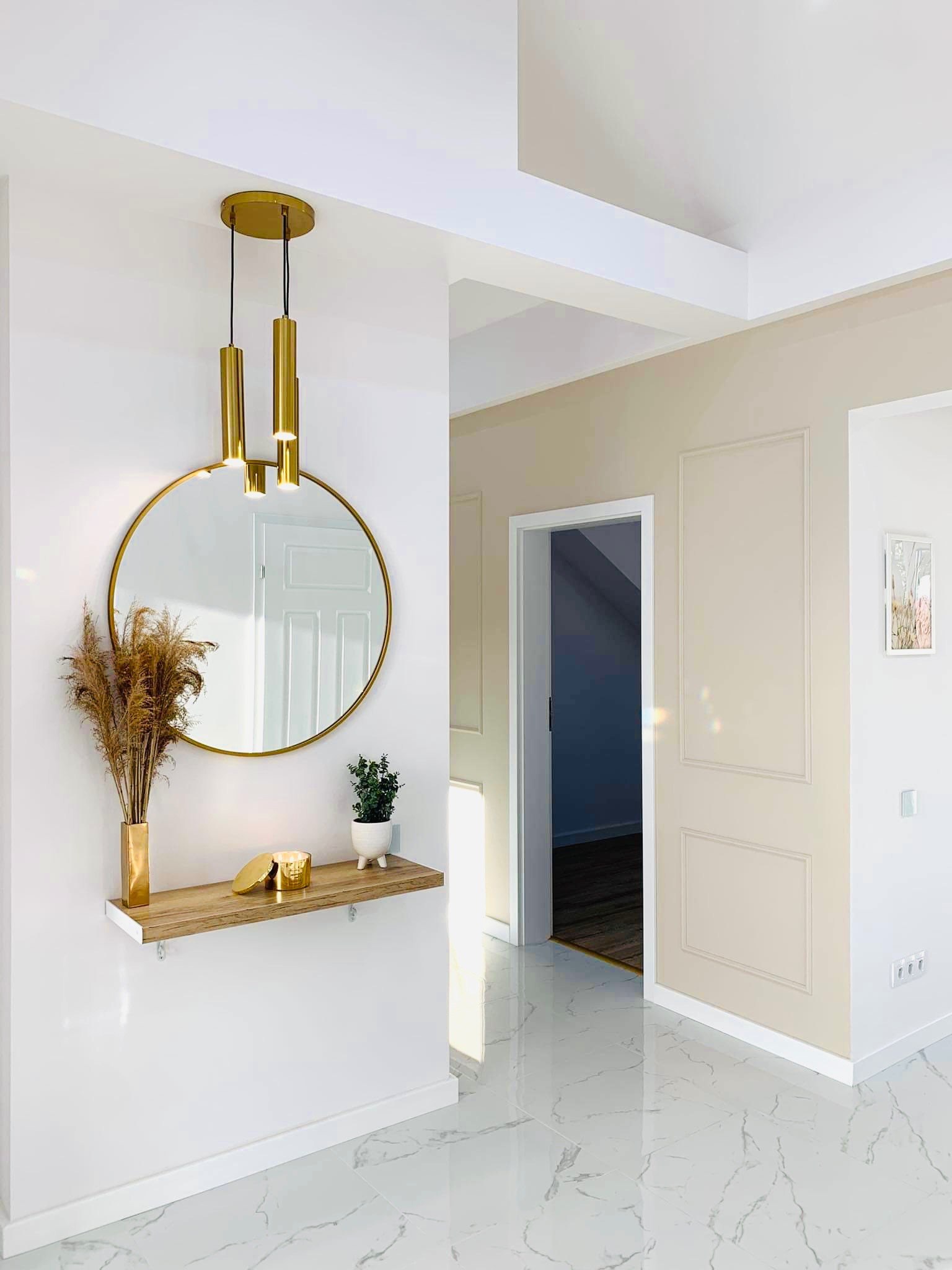 Nowoczesny przedpokój z okrągłym lustrem w złotej ramie, złotą lampą wiszącą, drewnianą półką z dekoracjami (wazon z suszonymi trawami, świeca w złotym pojemniku, roślina w doniczce) oraz marmurową...