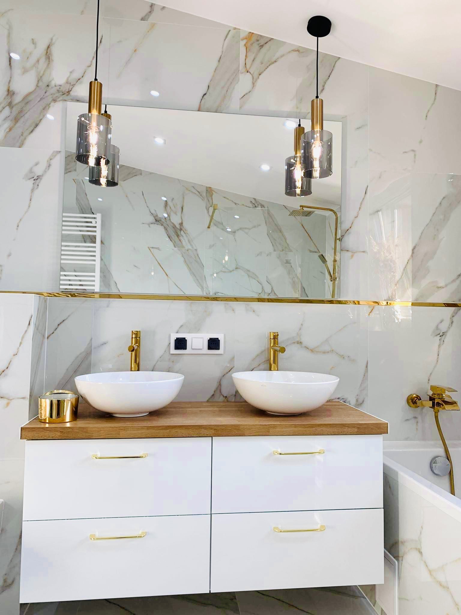 Nowoczesna łazienka z marmurowymi płytkami, dwiema białymi umywalkami nablatowymi z złotymi bateriami, drewnianym blatem, białymi szafkami z złotymi uchwytami i wiszącymi lampami o szklanych kloszach.