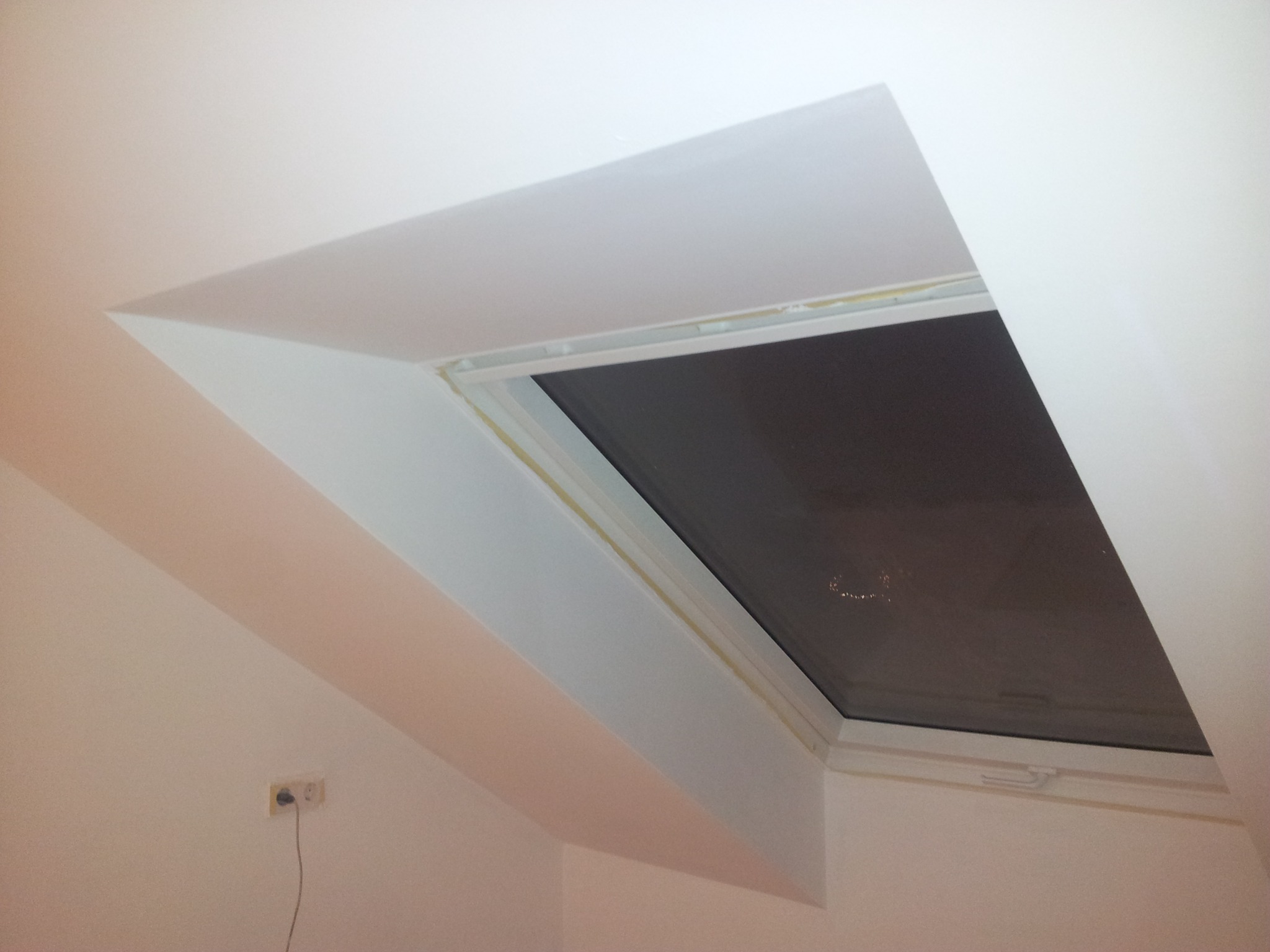 Poddasze z oknem dachowym po malowaniu, widoczne świeże białe ściany i sufit, silikonowa uszczelka wokół ramy okiennej, gniazdko elektryczne na ścianie.