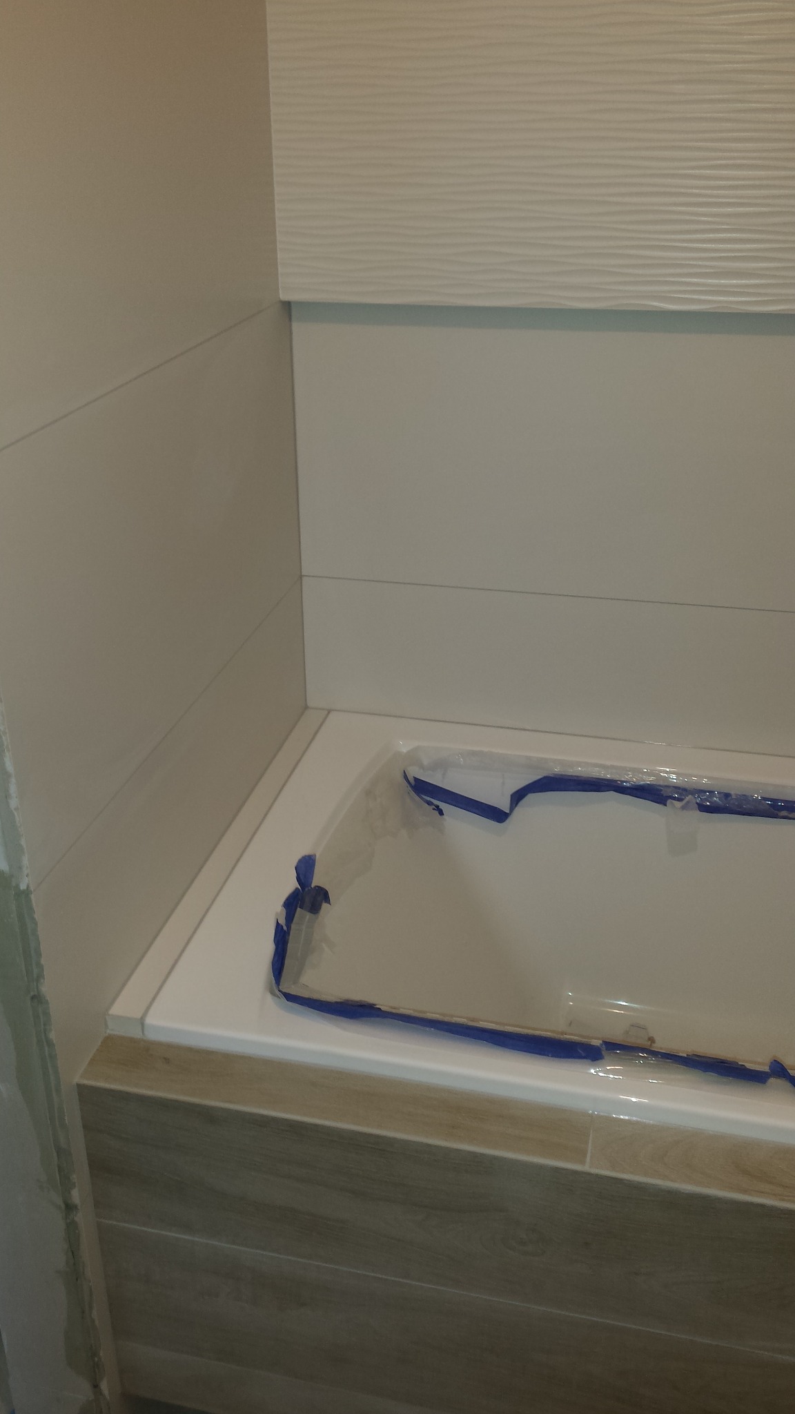 Wanna zabezpieczona folią malarską podczas prac wykończeniowych w łazience, widoczne płytki ścienne i obudowa wanny imitująca drewno.