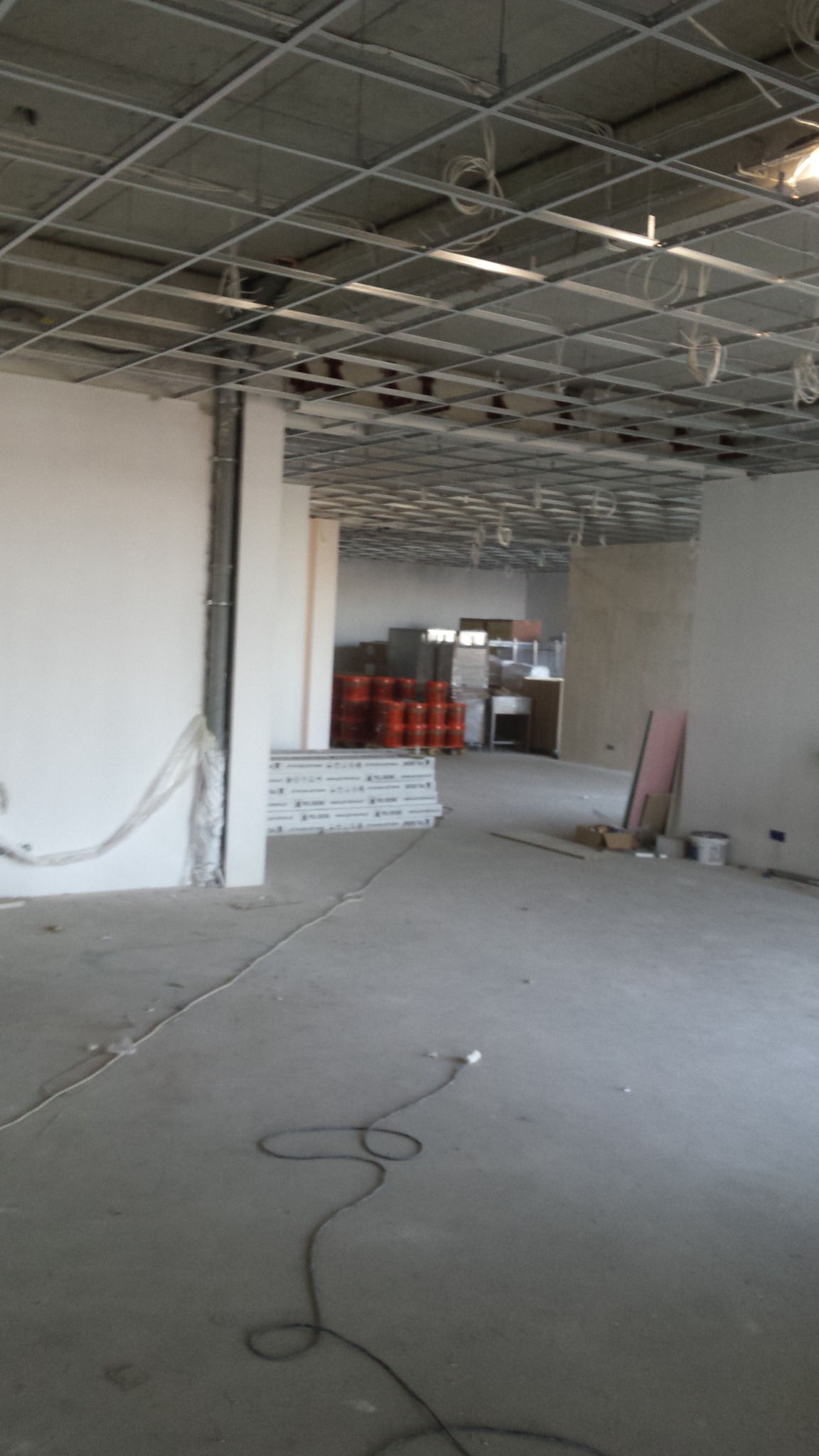 Wnętrze w trakcie prac wykończeniowych w Lublinie: widoczna surowa podłoga, ściany przygotowane do malowania, metalowa konstrukcja sufitu podwieszanego oraz materiały budowlane składowane w głębi...