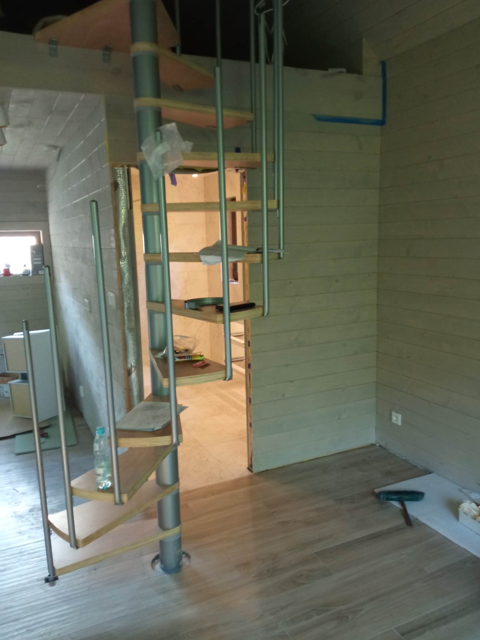 Wnętrze w trakcie wykańczania z drewnianymi panelami ściennymi, spiralne schody z butelką wody na jednym ze stopni, podłoga z paneli imitujących drewno i młotek leżący na podłodze.