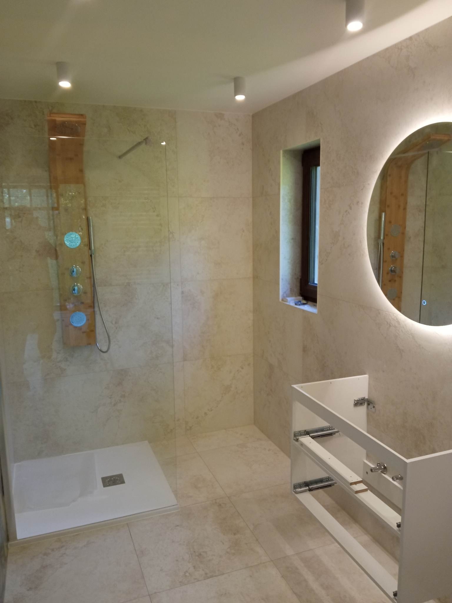 Wykończona łazienka z beżowymi płytkami, kabiną prysznicową typu walk-in z panelem drewnianym z dyszami, okrągłym lustrem z podświetleniem LED i szafką pod umywalkę bez frontów szuflad.