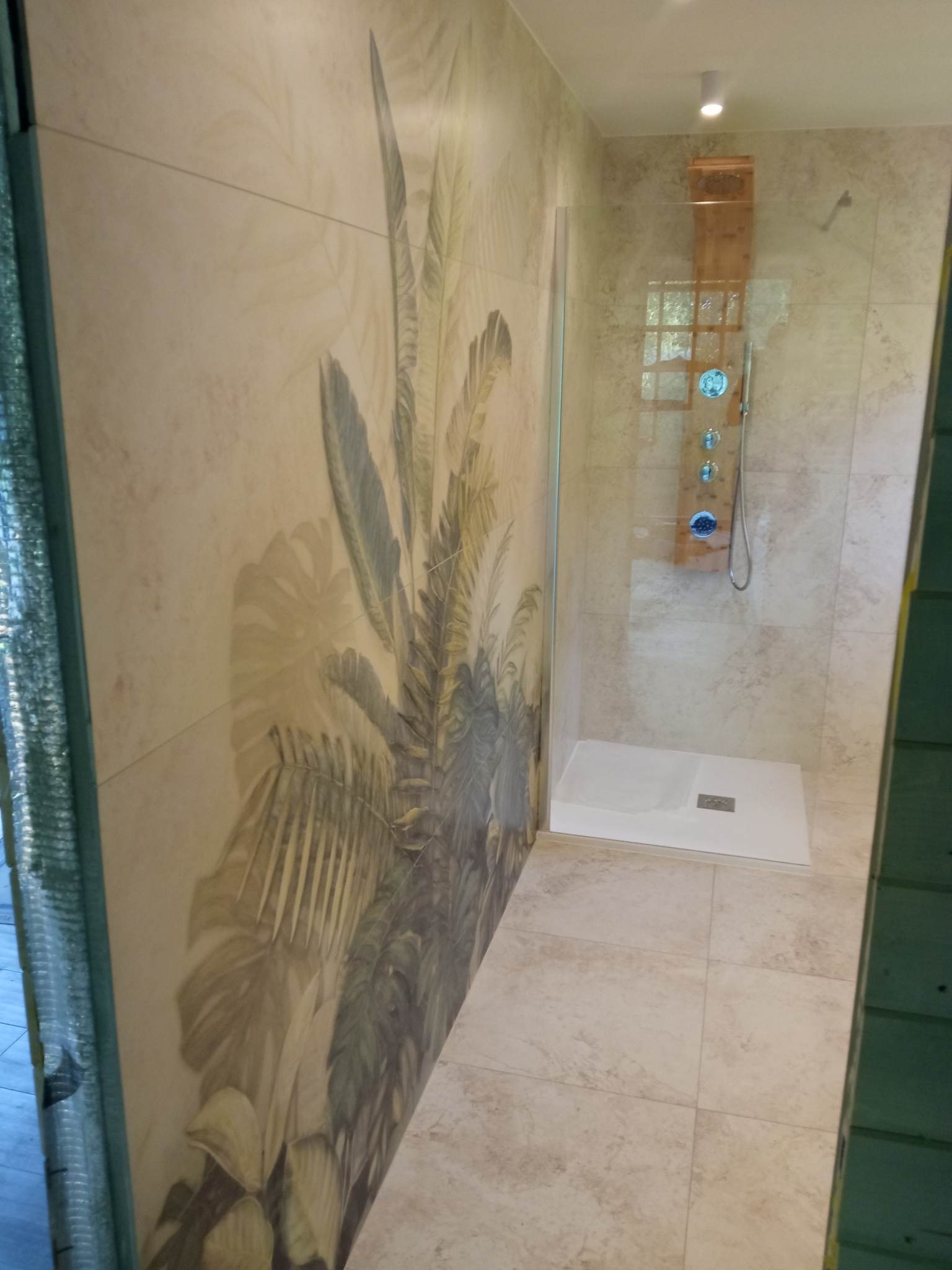 Nowoczesna kabina prysznicowa z panelem natryskowym i białym brodzikiem, ściana wyłożona płytkami z motywem tropikalnych liści, jasna podłoga z płytek imitujących kamień.