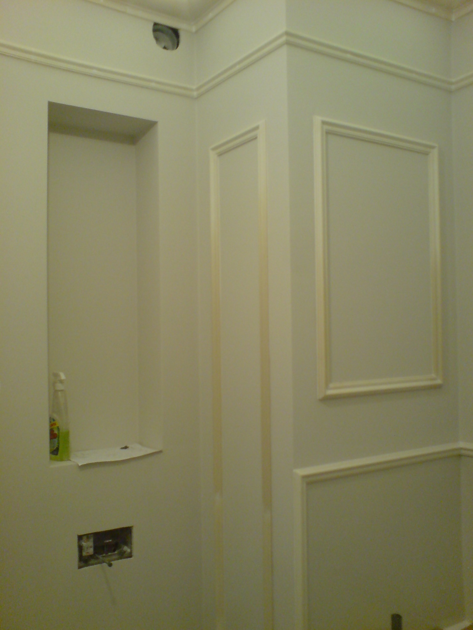 Ściana w trakcie prac wykończeniowych z dekoracyjnymi listwami, wnęką z półką i widocznym otworem instalacyjnym.