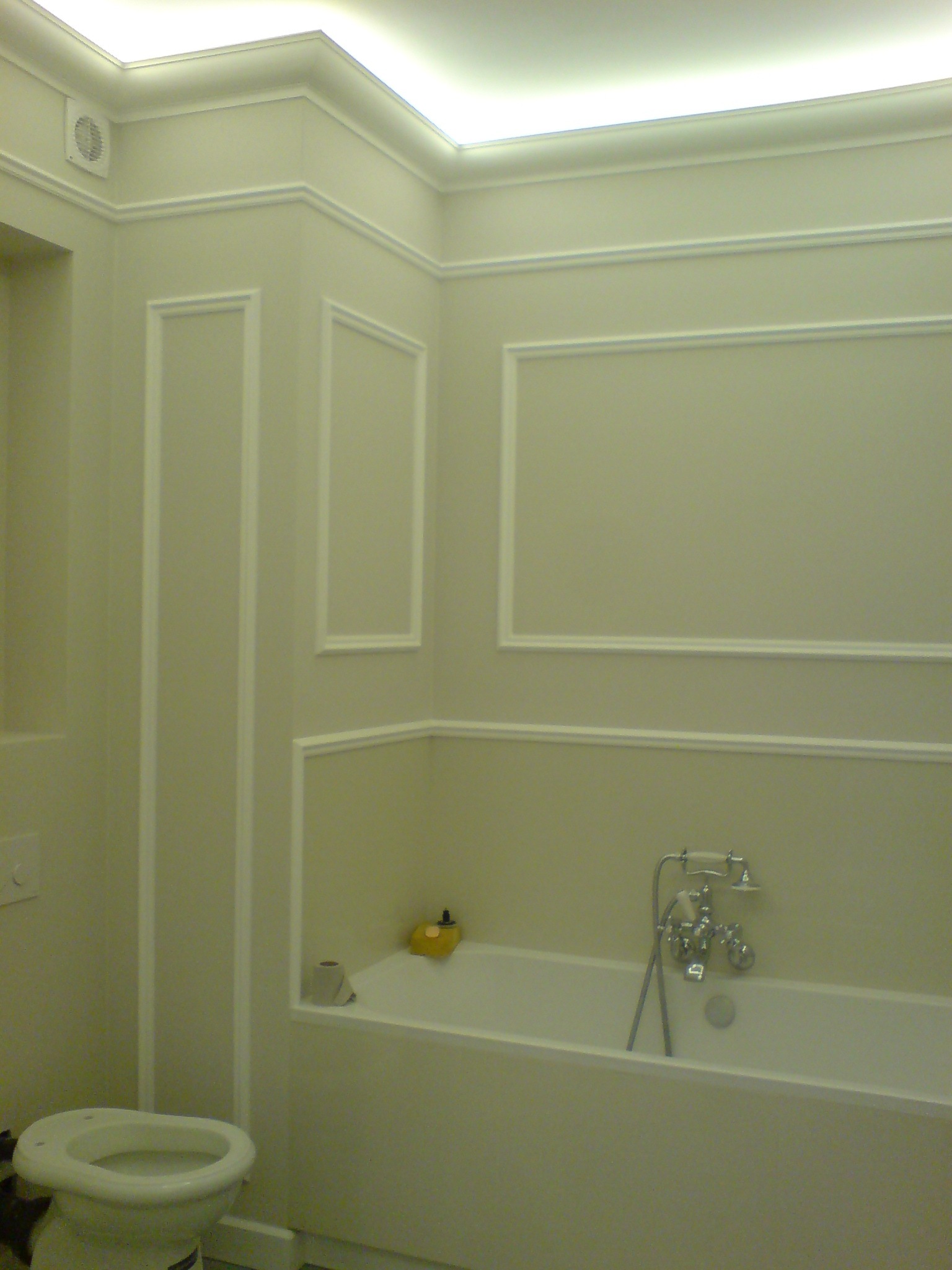 Wykończona łazienka w odcieniach beżu z białą armaturą, dekoracyjnymi listwami ściennymi i podświetlanym sufitem, widoczna toaleta i wanna z baterią.