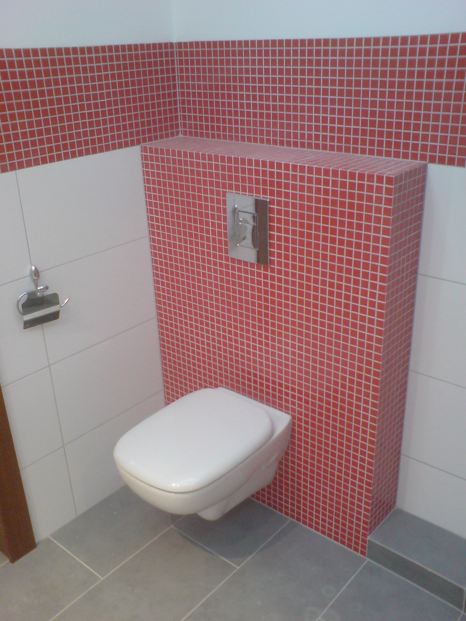 Wisząca toaleta z zabudową z czerwonej mozaiki, chromowany przycisk spłukiwania, białe i szare płytki na ścianach i podłodze, uchwyt na papier toaletowy.