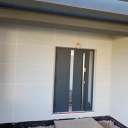 Ocieplona elewacja budynku z widoczną strukturą i podziałem na panele, grafitowe belki pod sufitem, nowoczesne drzwi wejściowe z przeszkleniami, widoczne elementy instalacji elektrycznej.