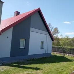 Dwukondygnacyjny dom z czerwoną dachówką i elewacją w odcieniach szarości i bieli, widoczny z perspektywy ogrodu, z fragmentem słupa energetycznego po lewej stronie.