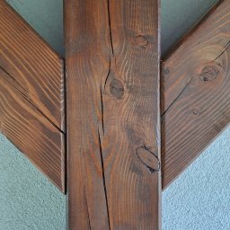 IVAN SHEVCHUK PRO100BUD - Fragment konstrukcji domu szkieletowego z brązowych belek drewnianych, widoczne sęki i pęknięcia drewna na tle jasnej, chropowatej ściany.