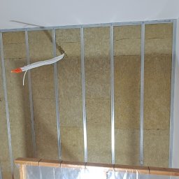 IVAN SHEVCHUK PRO100BUD - Ściana w trakcie ocieplania wełną mineralną, widoczne profile metalowe, przewody elektryczne zabezpieczone peszlem z pomarańczowymi końcówkami, fragment ościeżnicy drzwiowej zabezpieczonej folią.