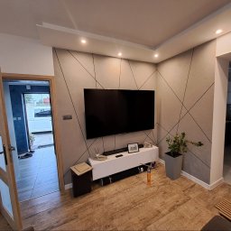 IVAN SHEVCHUK PRO100BUD - Nowoczesny salon z szarą ścianą zdobioną geometrycznym wzorem, drewnianą podłogą i białymi meblami RTV. W tle widoczny fragment kuchni i otwarte drzwi z widokiem na podjazd.