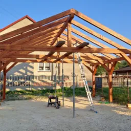 Drewniana konstrukcja zadaszenia w trakcie budowy, widoczne słupy i krokwie, drabina oparta o konstrukcję, w tle budynki mieszkalne.